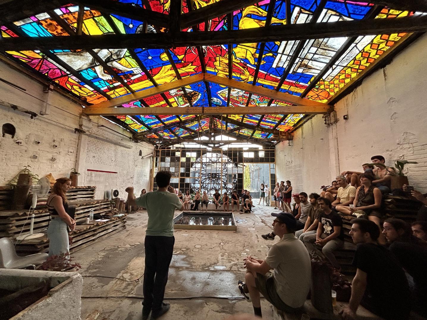People sitting in an old empty stable like building with a stained galss roof, listening to someone speak