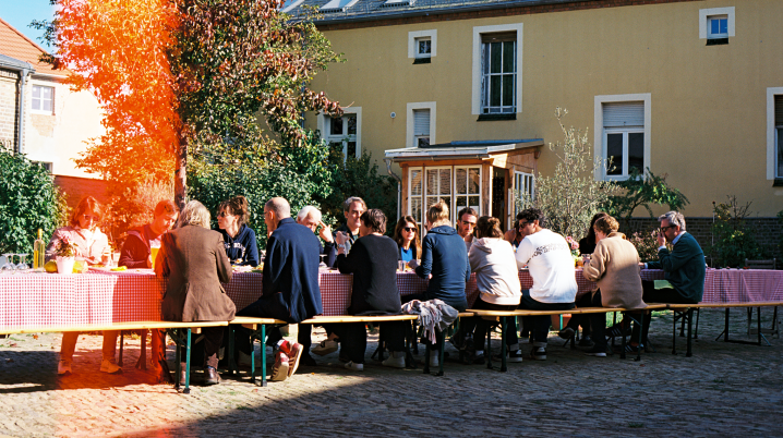 A group of people sitting at a large table outside having a lunch, in front of a yellow building 